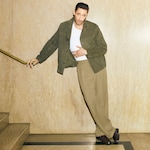 Placeholder for: Man in a brown jacket and beige pants leans casually against a wall, holding a stair railing.