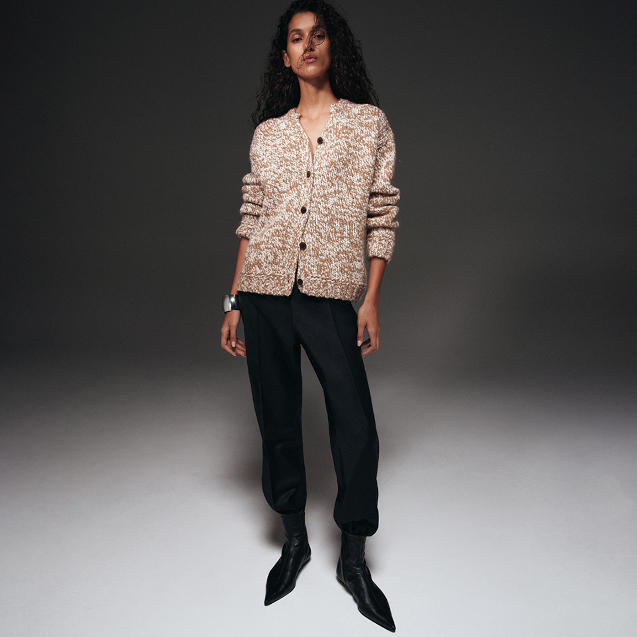 Woman wearing a textured brown cardigan, black pants, and pointed black boots, standing against a plain dark background.