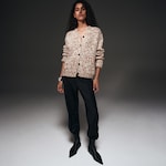Placeholder for: Woman wearing a textured brown cardigan, black pants, and pointed black boots, standing against a plain dark background.