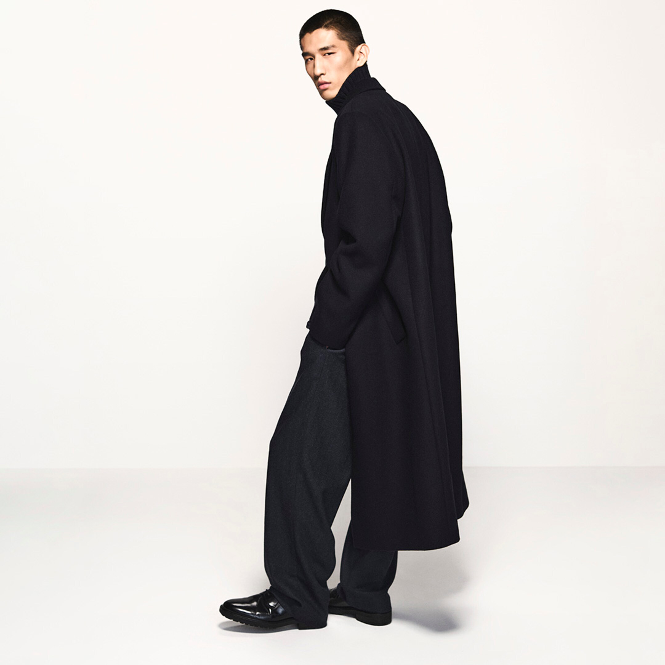 Man in a long black coat and turtleneck, with dark trousers and shiny black shoes, walking stylishly against a plain background.