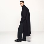 Placeholder for: Man in a long black coat and turtleneck, with dark trousers and shiny black shoes, walking stylishly against a plain background.