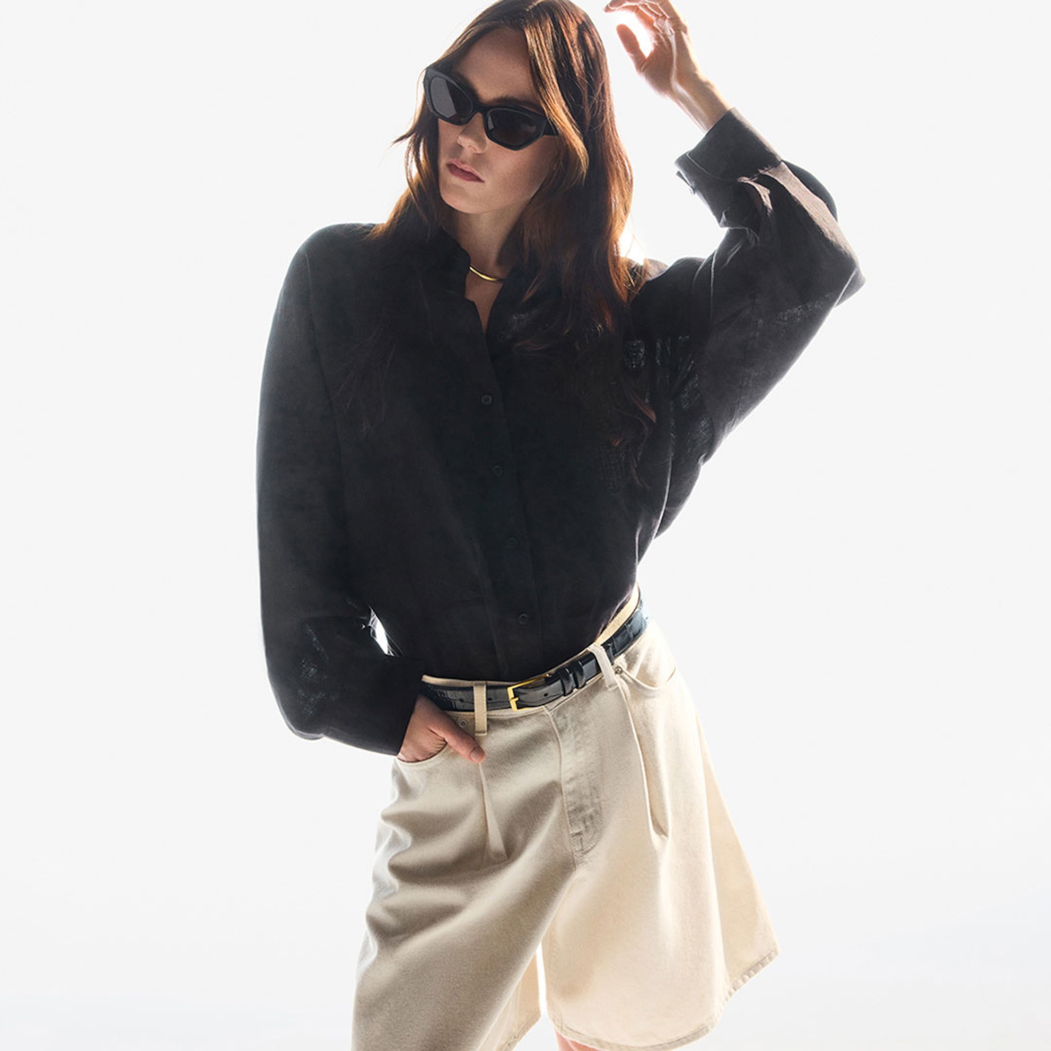 Woman in sunglasses wearing a black shirt and beige shorts poses confidently against a white background.