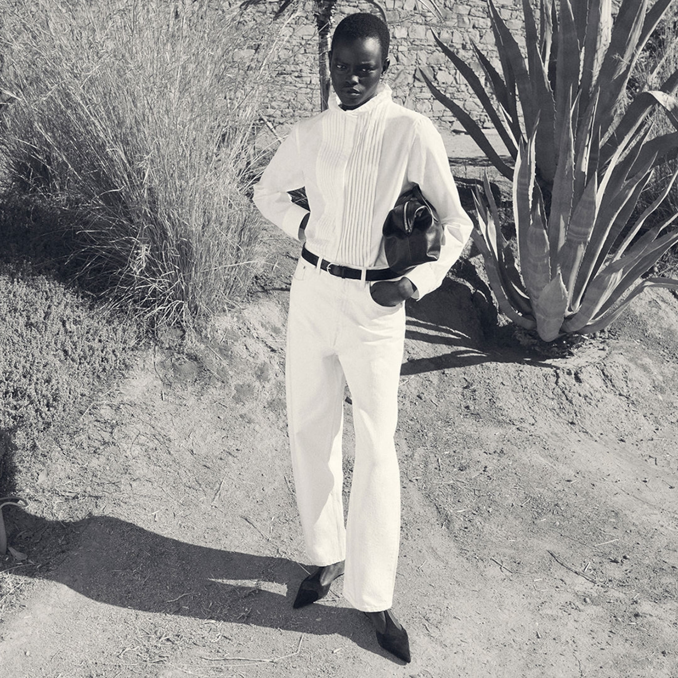 Person in a white outfit standing confidently in a desert setting with plants and a stone wall in the background.