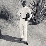 Placeholder for: Person in a white outfit standing confidently in a desert setting with plants and a stone wall in the background.