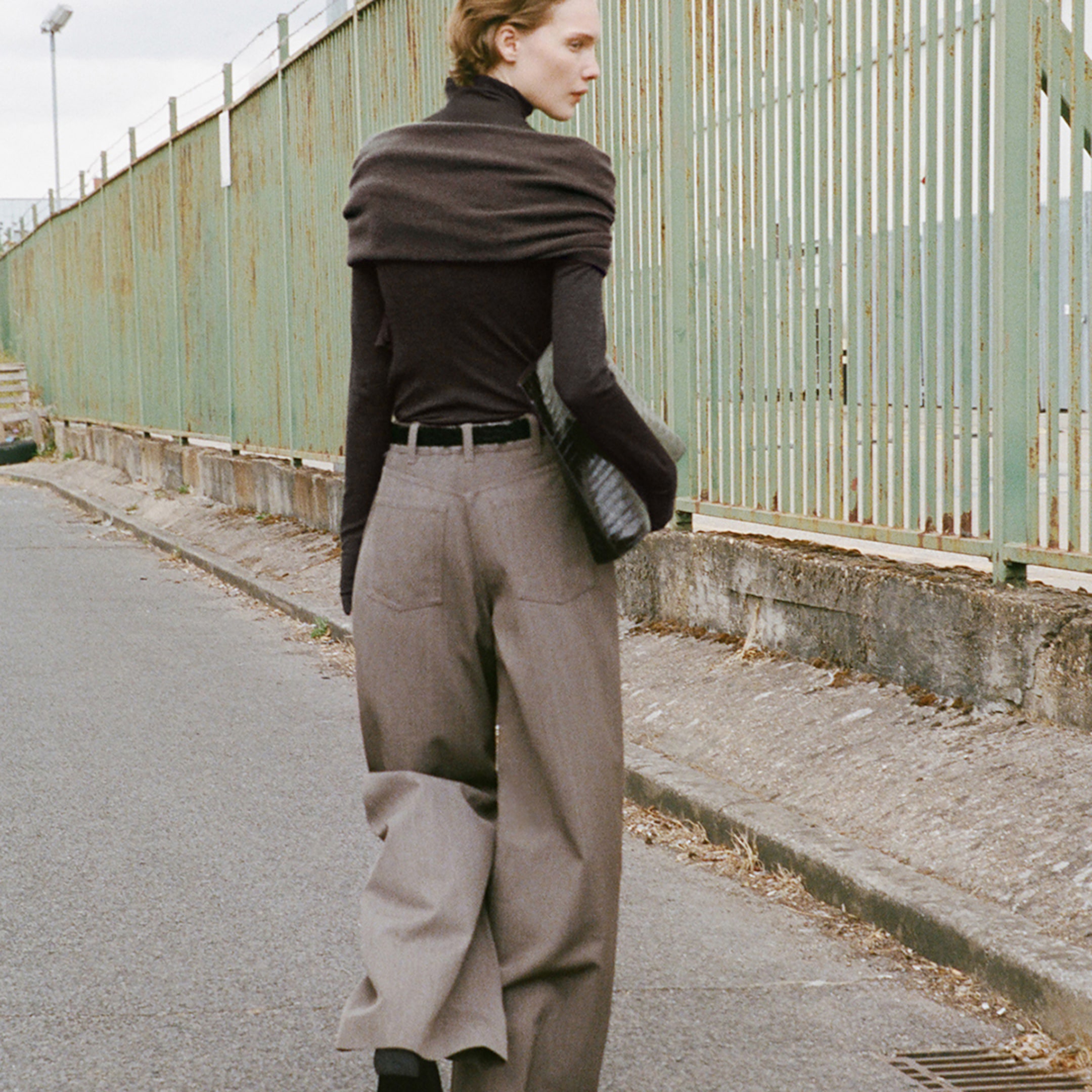 Person in stylish, loose-fitting gray trousers and a dark top walks on a paved path beside a green metal fence, holding a bag.
