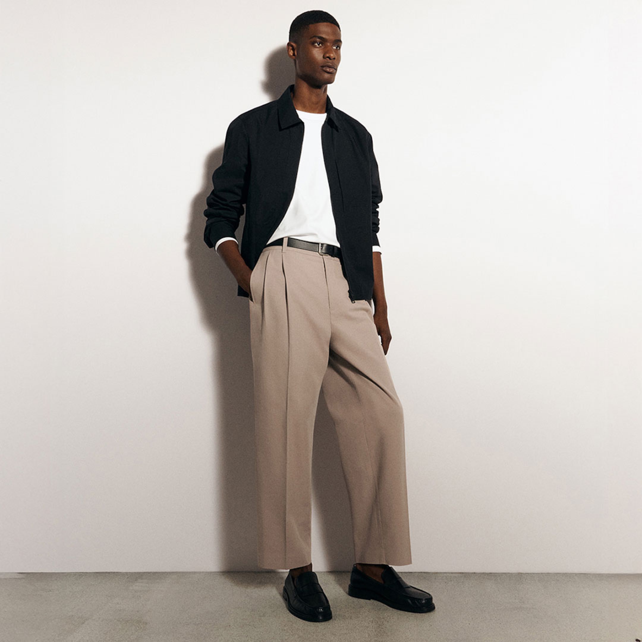 A man wearing a black jacket, white shirt, beige trousers, and black loafers stands against a white wall with one hand in his pocket.