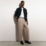 Placeholder for: A man wearing a black jacket, white shirt, beige trousers, and black loafers stands against a white wall with one hand in his pocket.