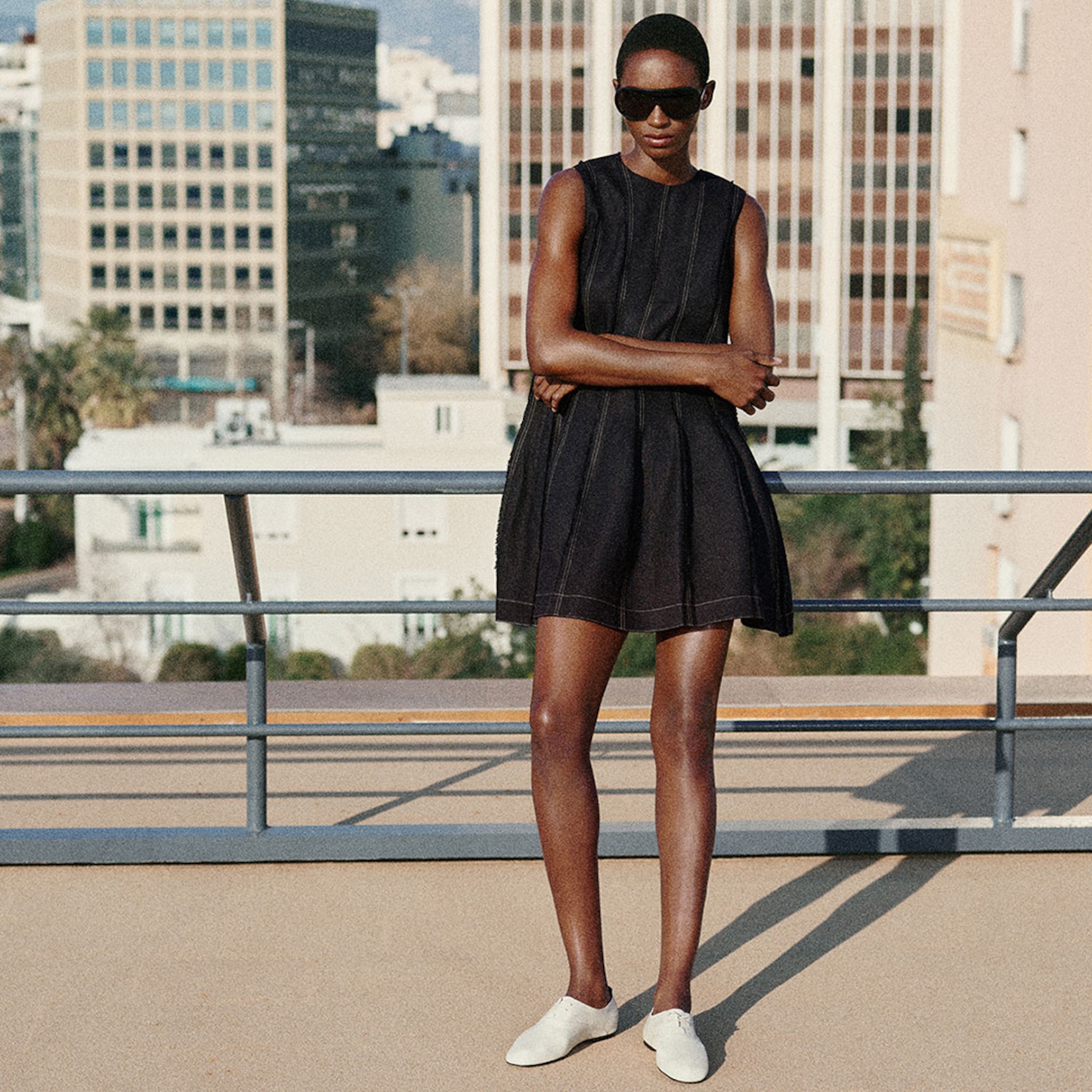 Person wearing sunglasses, a sleeveless black dress, and white shoes standing on a rooftop with city buildings in the background.