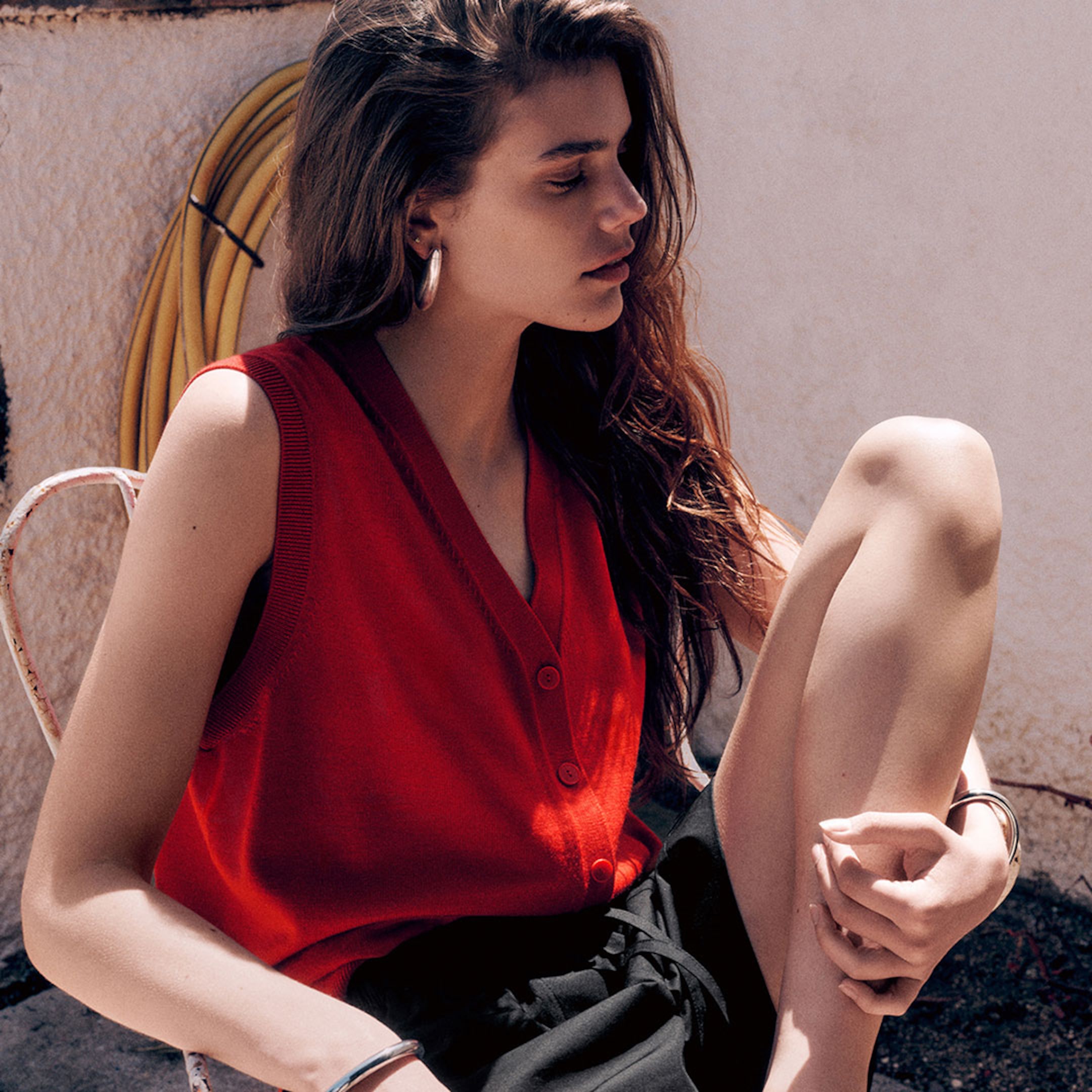 Woman in a red sleeveless top, sitting with eyes closed, her long hair cascading over her shoulders. She wears hoop earrings.