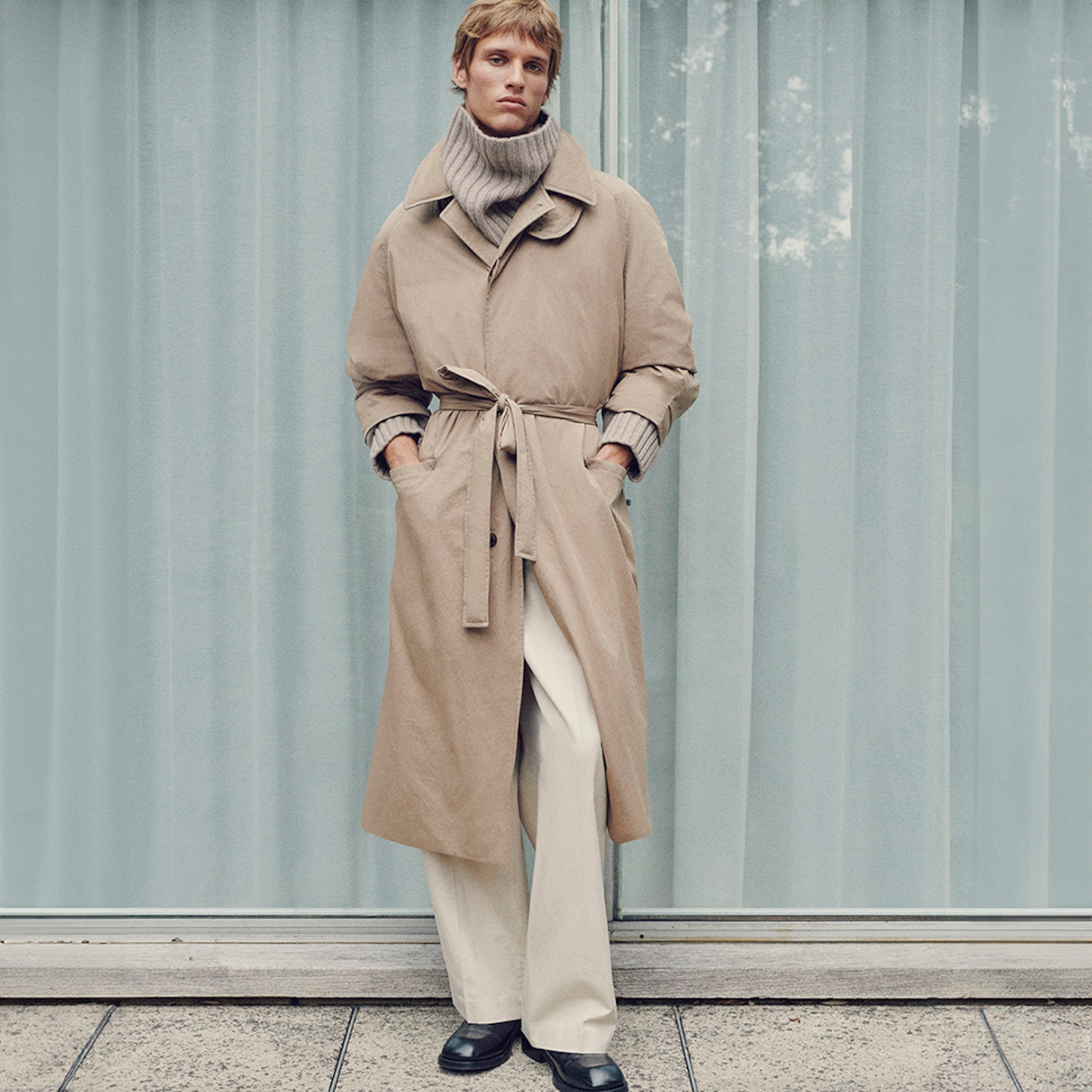 Person wearing a beige trench coat with a belt, gray turtleneck, white pants, and black shoes, standing in front of a curtain.