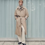 Placeholder for: Person wearing a beige trench coat with a belt, gray turtleneck, white pants, and black shoes, standing in front of a curtain.