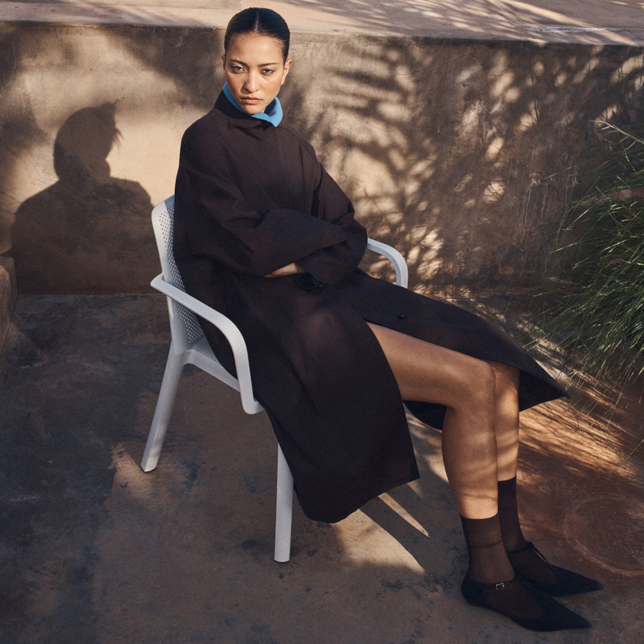 Person sitting on a white chair, wearing a dark coat with blue collar, in a sunlit outdoor setting with a stone wall and shadow patterns visible.