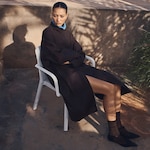 Placeholder for: Person sitting on a white chair, wearing a dark coat with blue collar, in a sunlit outdoor setting with a stone wall and shadow patterns visible.
