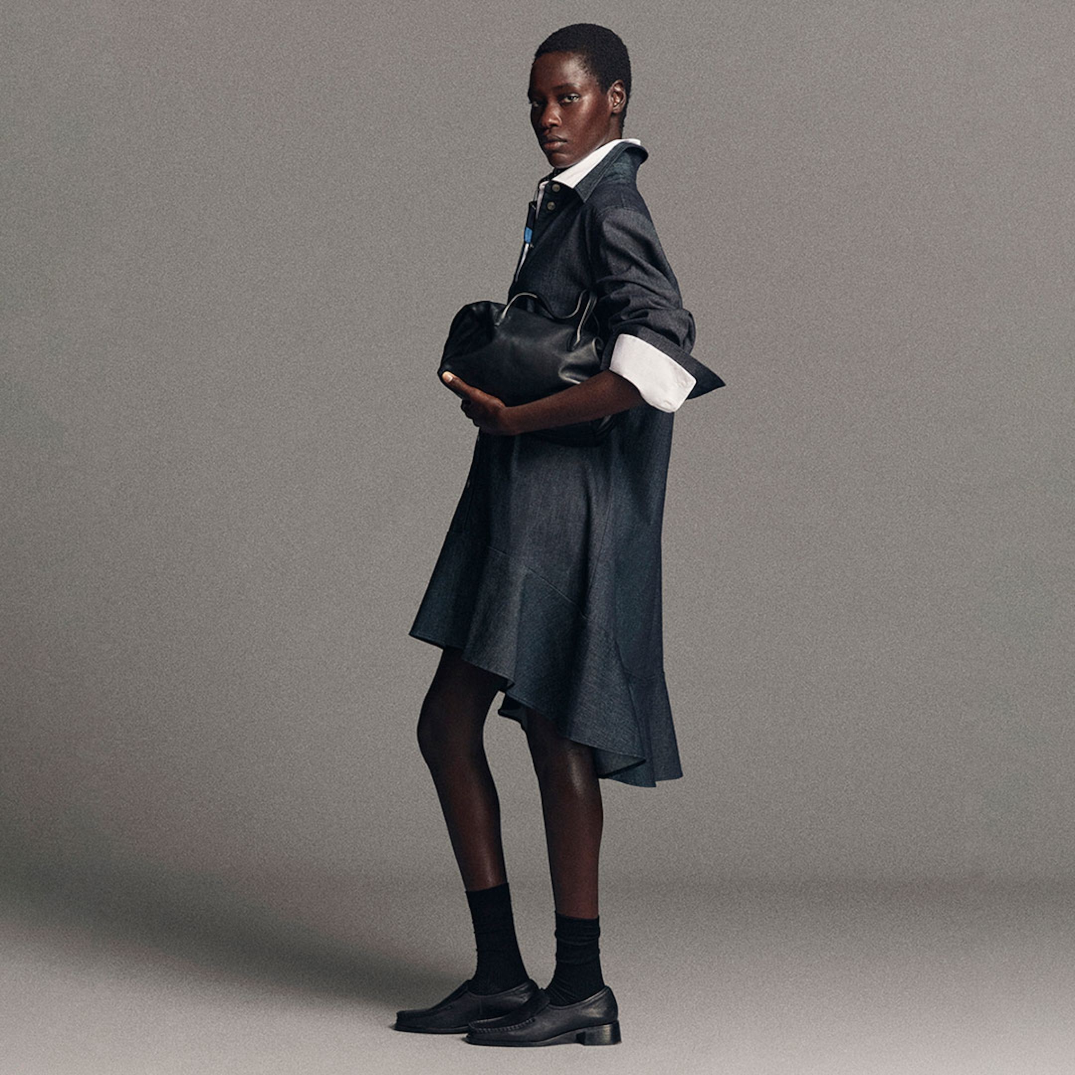 A person in a dark shirt dress and white collar holds a black handbag. They wear black socks and shoes, standing against a gray background.