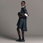 Placeholder for: A person in a dark shirt dress and white collar holds a black handbag. They wear black socks and shoes, standing against a gray background.