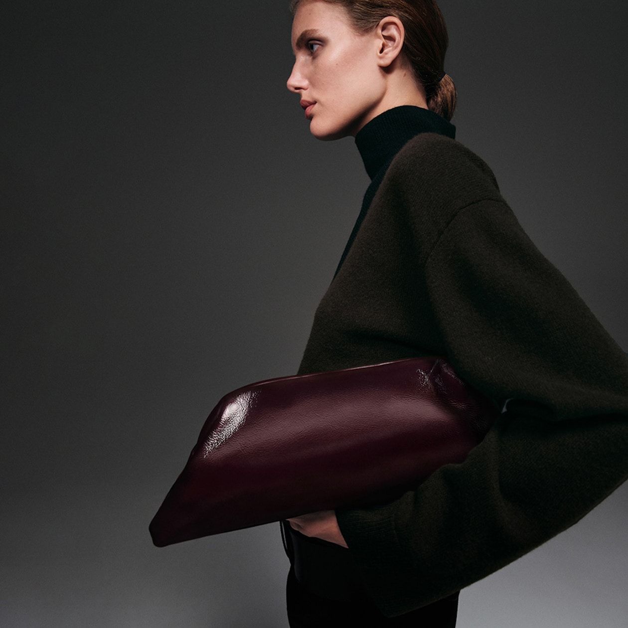 Person in a dark sweater holding a large burgundy leather bag under their arm, with a side profile against a gray background.