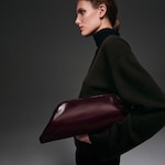 Placeholder for: Person in a dark sweater holding a large burgundy leather bag under their arm, with a side profile against a gray background.