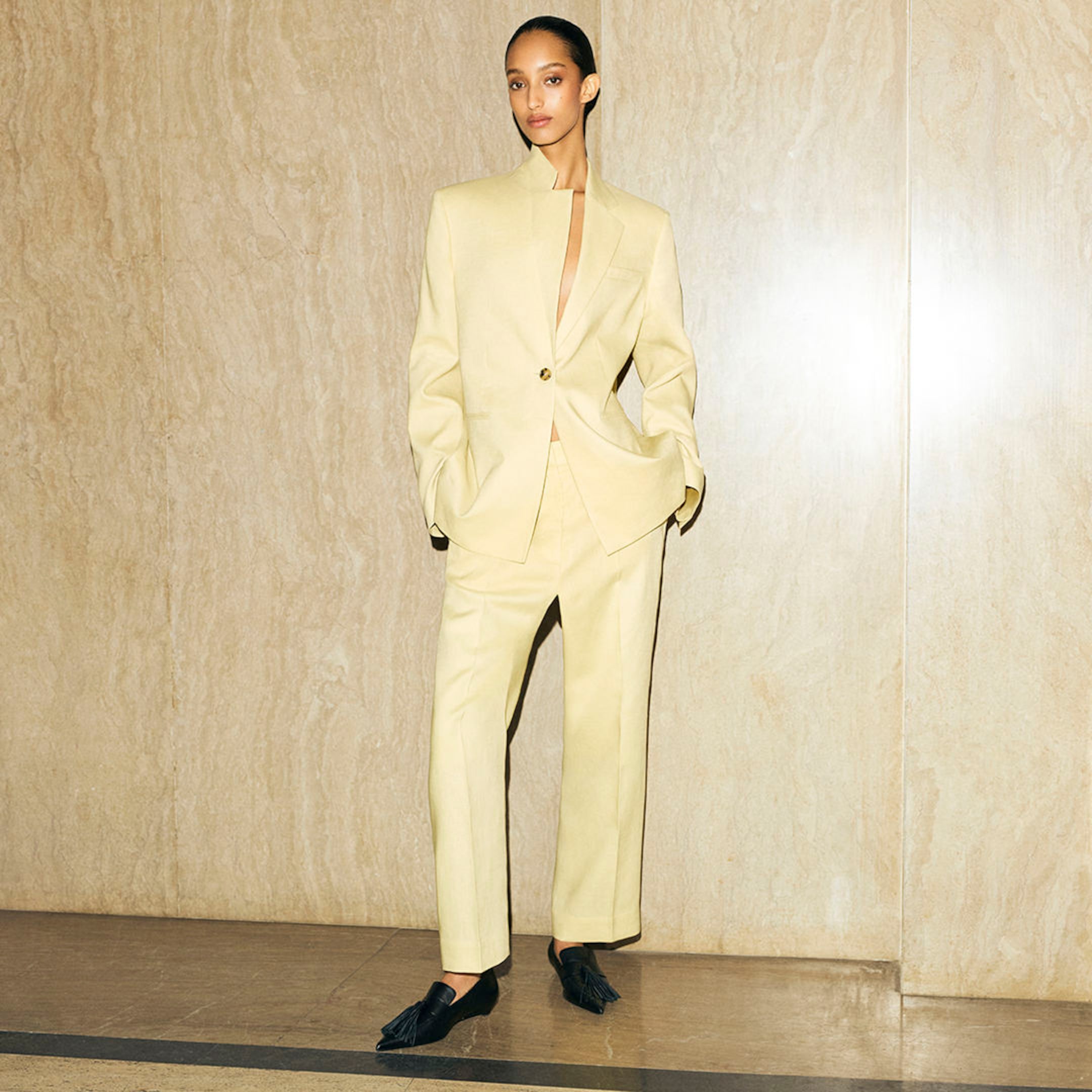 Person wearing a light beige suit with hands in pockets, standing against a marble wall, and wearing black shoes.