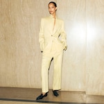 Placeholder for: Person wearing a light beige suit with hands in pockets, standing against a marble wall, and wearing black shoes.