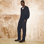 Placeholder for: Man in a navy cardigan and trousers stands against a textured beige marble wall, hands in pockets, wearing a white shirt and dark shoes.