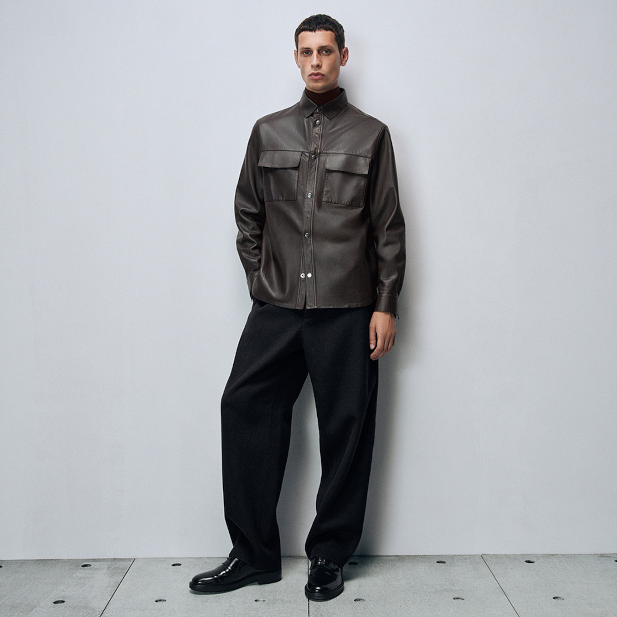 Person in a dark brown leather jacket and black pants stands against a plain gray background, hands in pockets, gazing forward.