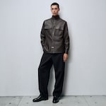 Placeholder for: Person in a dark brown leather jacket and black pants stands against a plain gray background, hands in pockets, gazing forward.