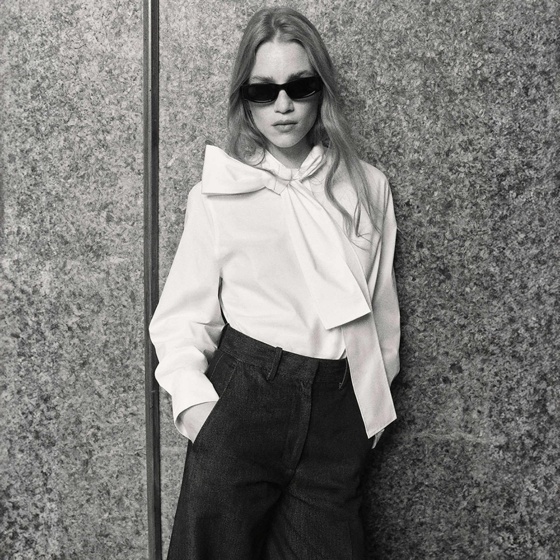 Woman with long hair and sunglasses wearing a white blouse with a large bow and high-waisted pants, standing against a textured wall. Black and white photo.