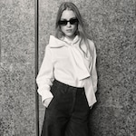 Placeholder for: Woman with long hair and sunglasses wearing a white blouse with a large bow and high-waisted pants, standing against a textured wall. Black and white photo.