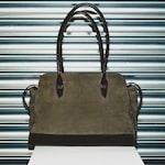 Placeholder for: A stylish suede handbag with dark leather handles and accents, displayed against a metal shutter background on a white pedestal.