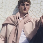 Placeholder for: Person wearing a pink shirt with a sweater draped over their shoulders, against a textured, geometric white wall.