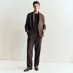 Placeholder for: Man wearing a brown suit with a collarless blazer and matching pants, hands in pockets, standing against a light background.