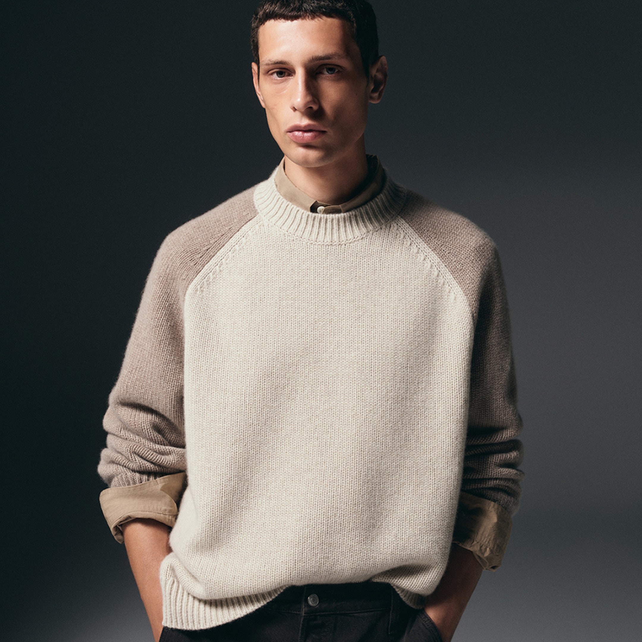 Man wearing a beige knit sweater over a collared shirt, with one hand in pocket against a dark background.