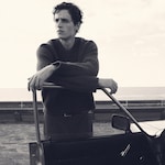 Placeholder for: Man in a sweater leans on car door, gazing thoughtfully into the distance. Black and white photo creates a vintage, contemplative mood.