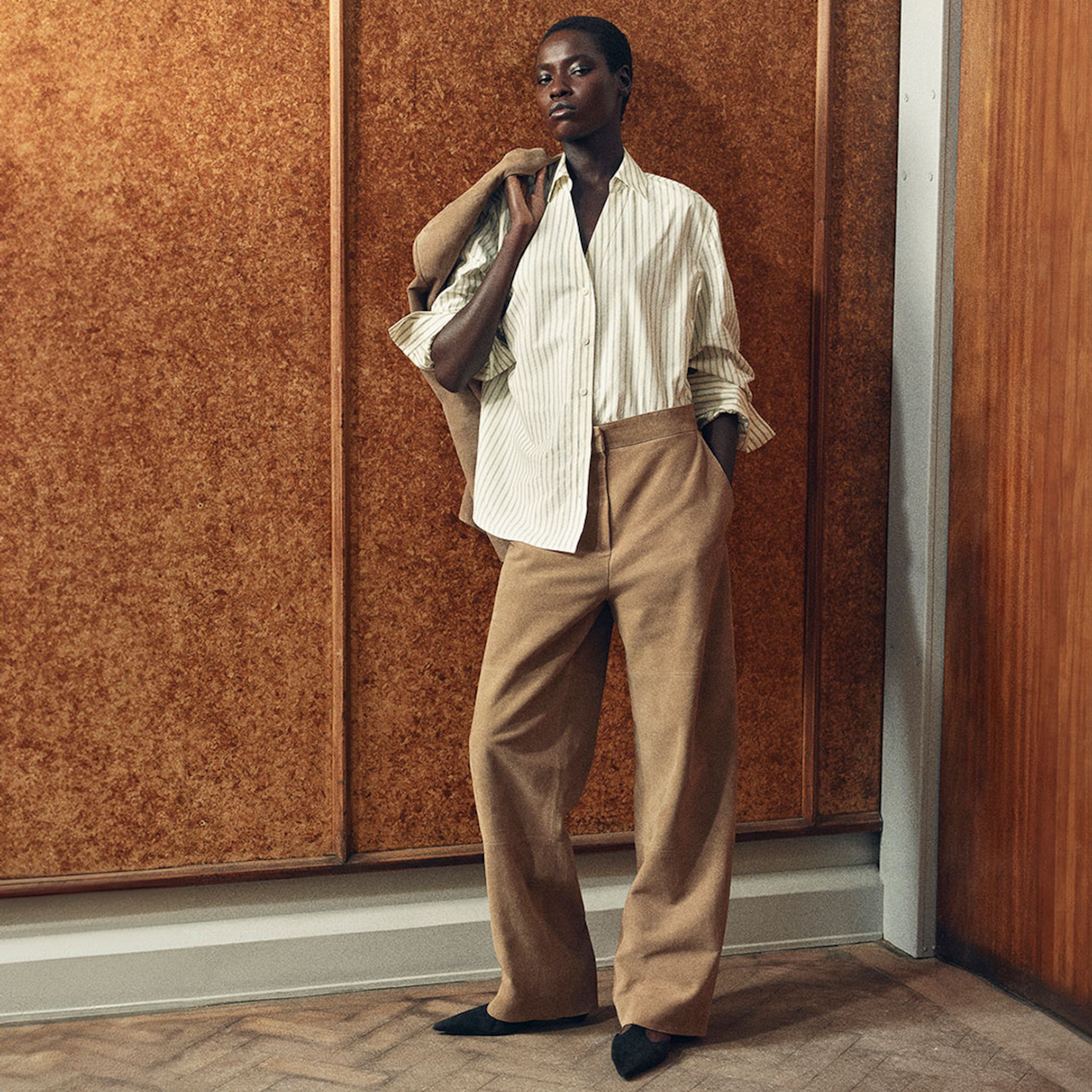 Model in a striped shirt and tan pants, holding a matching jacket. The background is a textured wooden wall. They exude confidence and poise.