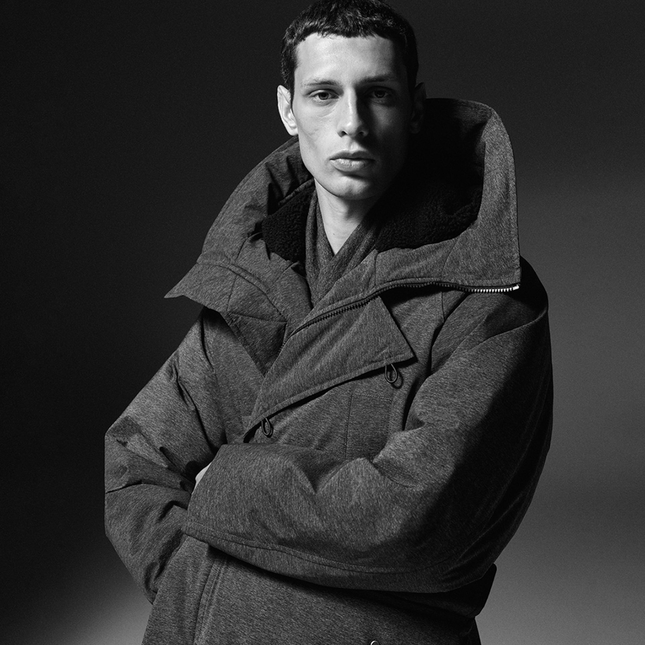Black and white portrait of a person wearing a thick, textured hooded jacket, looking directly at the camera against a dark background.