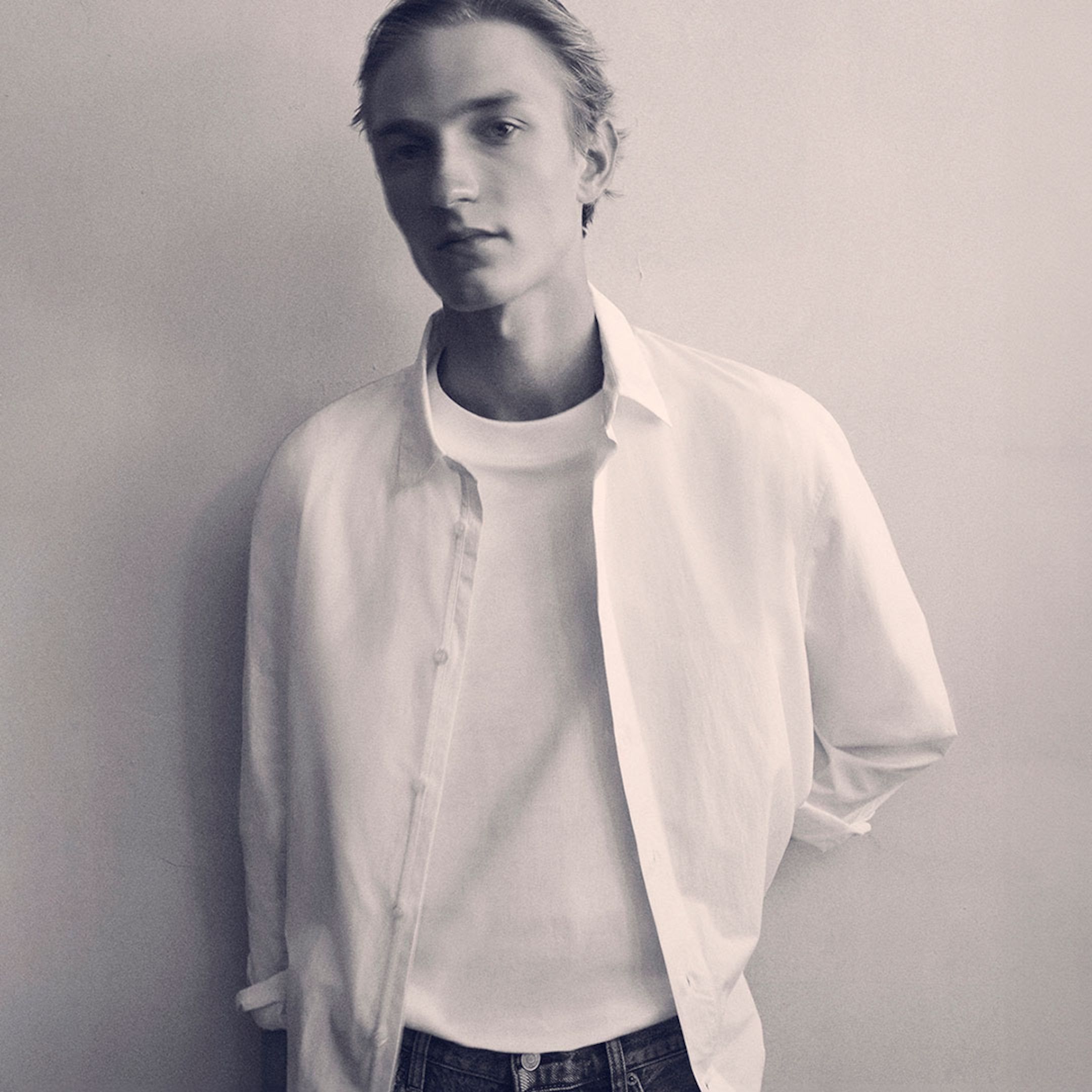 Black and white photo of a person with short hair, wearing a white shirt over a white tee, standing against a neutral background.