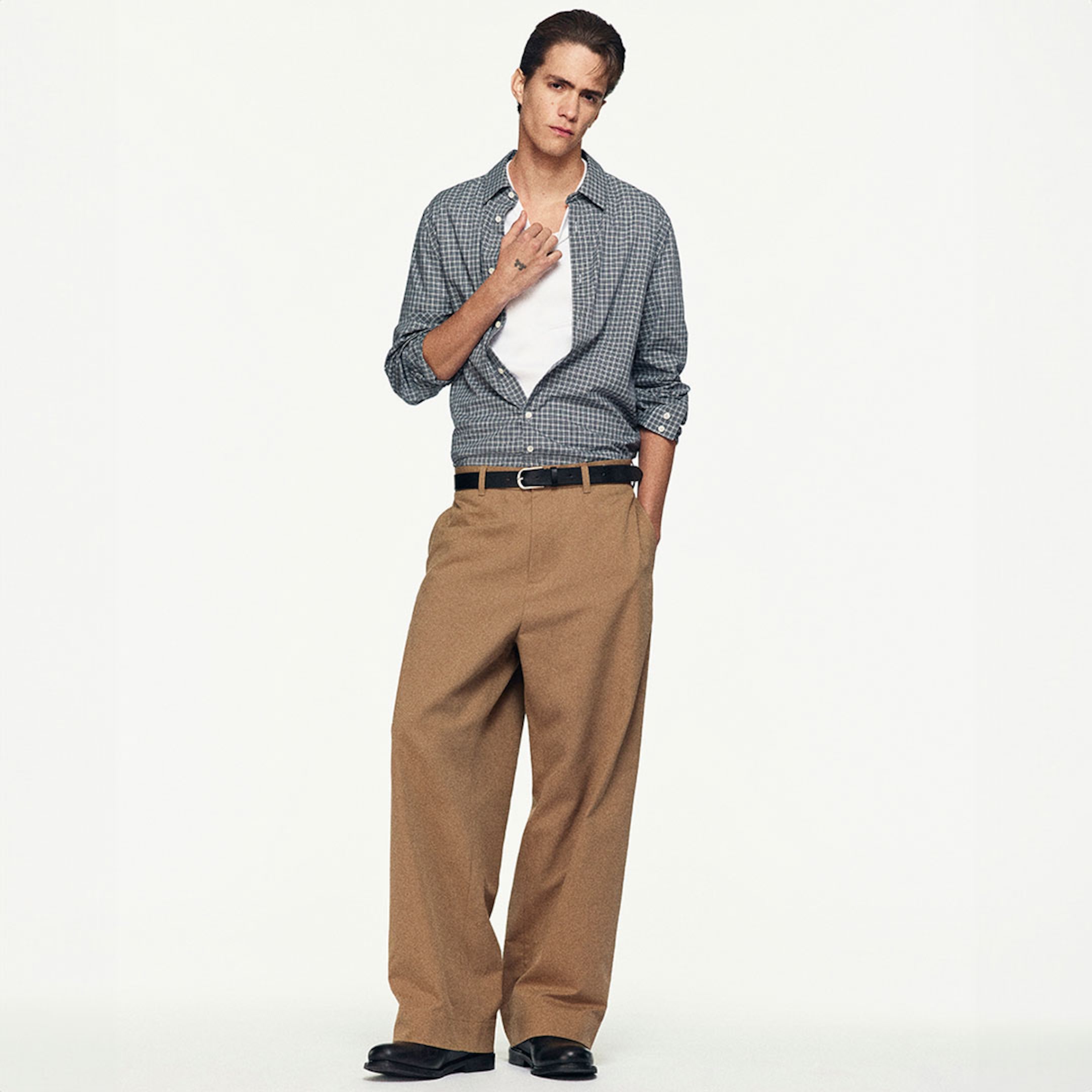 Model wearing a checked shirt over a white tee, brown wide-leg trousers, and black shoes, posing with one hand in the pocket.