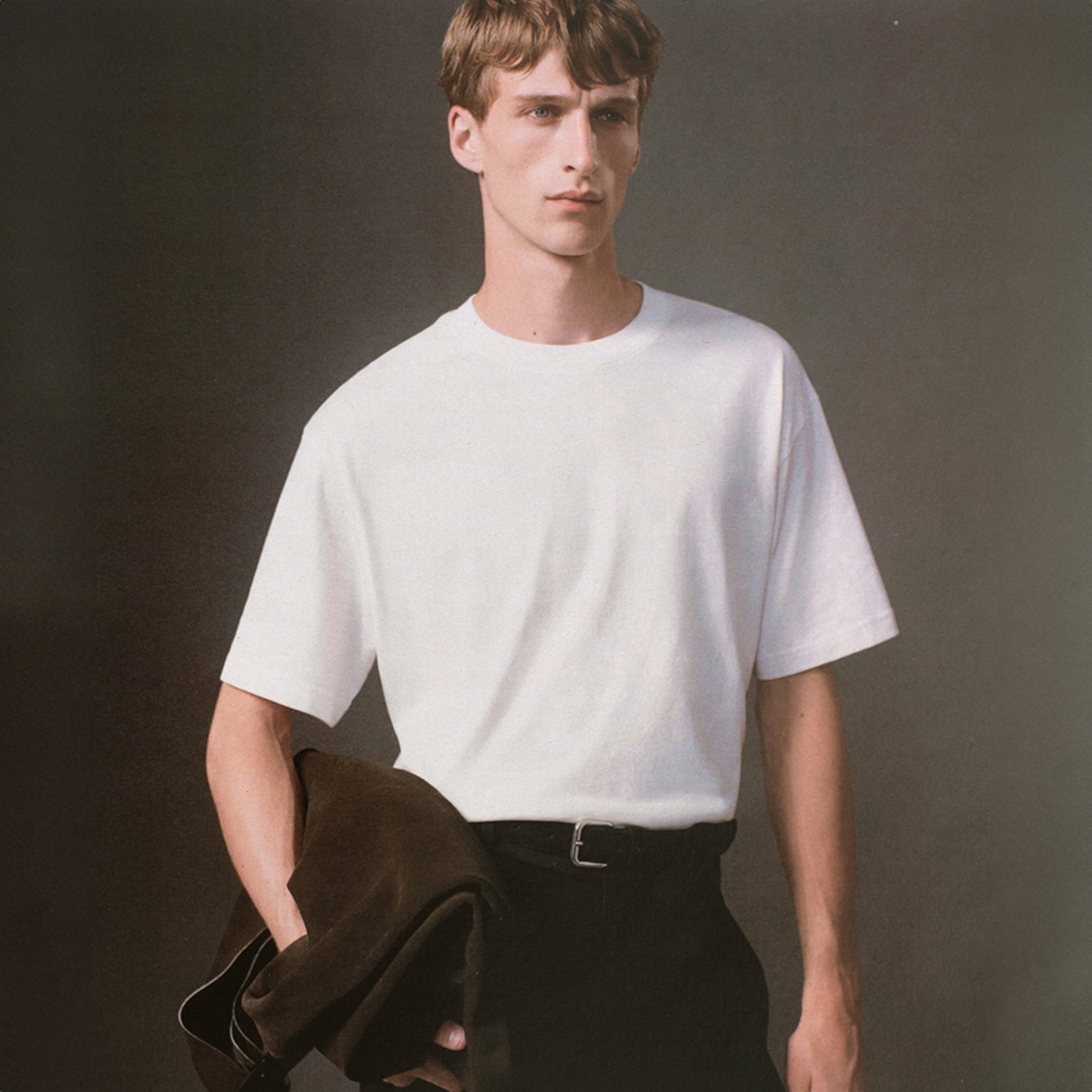 A person wearing a plain white T-shirt and holding a dark jacket, set against a neutral gray background.