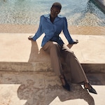 Placeholder for: Person sitting on a stone ledge by a pool, wearing a blue blouse and brown trousers, with chic black shoes.