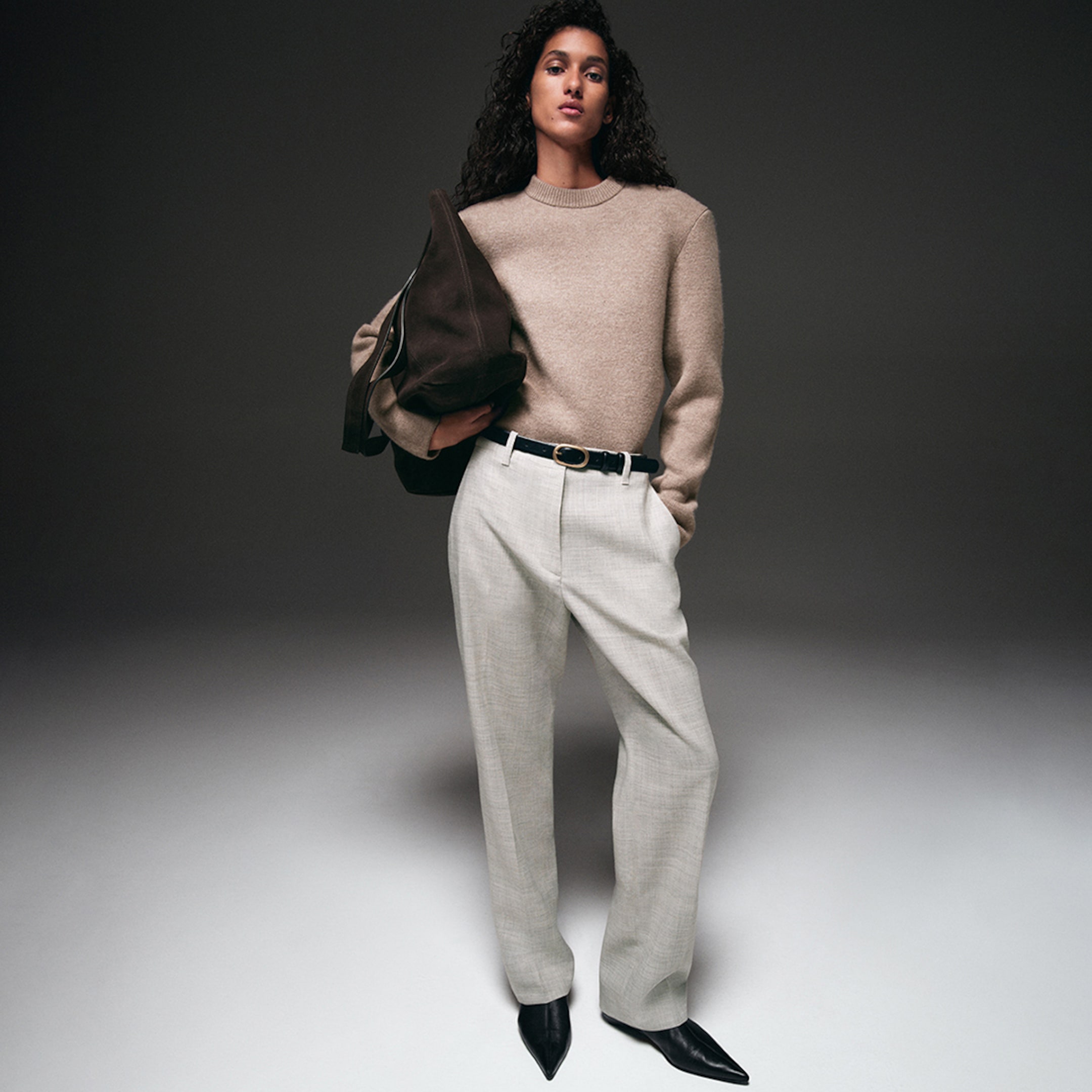 A person wearing a beige sweater, gray trousers, and black pointed shoes, holding a brown bag, standing against a neutral background.