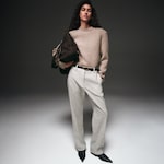 Placeholder for: A person wearing a beige sweater, gray trousers, and black pointed shoes, holding a brown bag, standing against a neutral background.