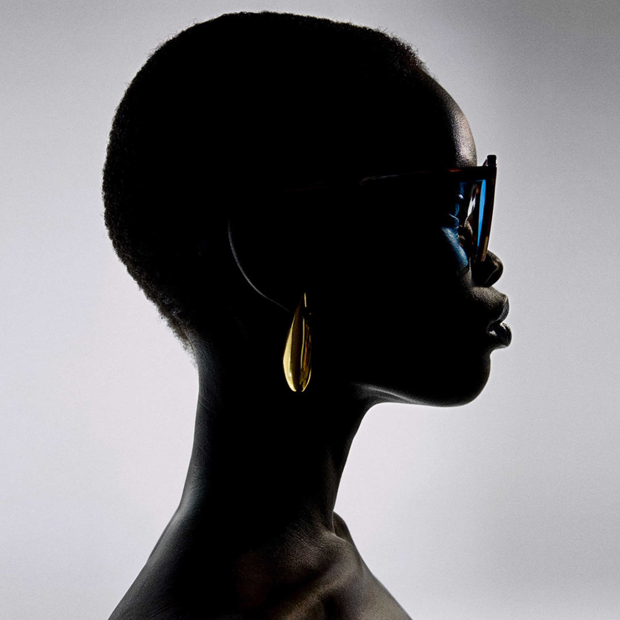 Silhouette of a person in profile wearing large sunglasses and a gold earring, set against a light gray background.