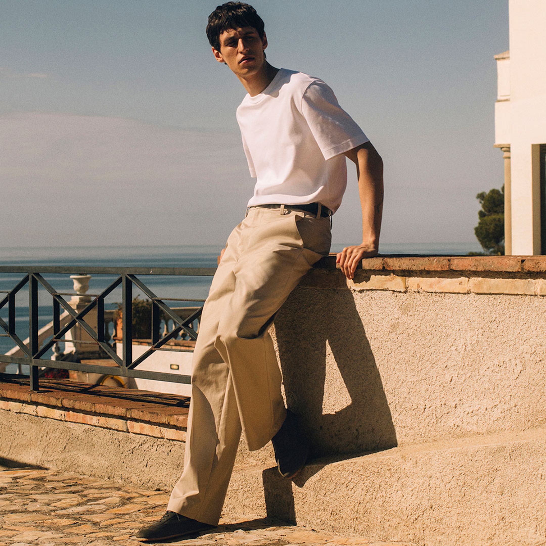 A full-length lifestyle photograph of a man leaning back against a sunlit stone wall on an outdoor terrace. He is wearing a crisp white short-sleeved T-shirt tucked into beige tailored trousers with a slim dark belt. The look is finished with dark low-top sneakers. The background features a black metal railing and a serene view of the sea under a clear sky.