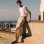 Placeholder for: A full-length lifestyle photograph of a man leaning back against a sunlit stone wall on an outdoor terrace. He is wearing a crisp white short-sleeved T-shirt tucked into beige tailored trousers with a slim dark belt. The look is finished with dark low-top sneakers. The background features a black metal railing and a serene view of the sea under a clear sky.
