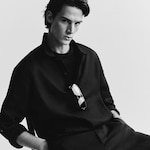 Placeholder for: Black and white portrait of a person wearing a dark shirt with sunglasses hanging from the collar, sitting on a chair with one hand in their pocket.