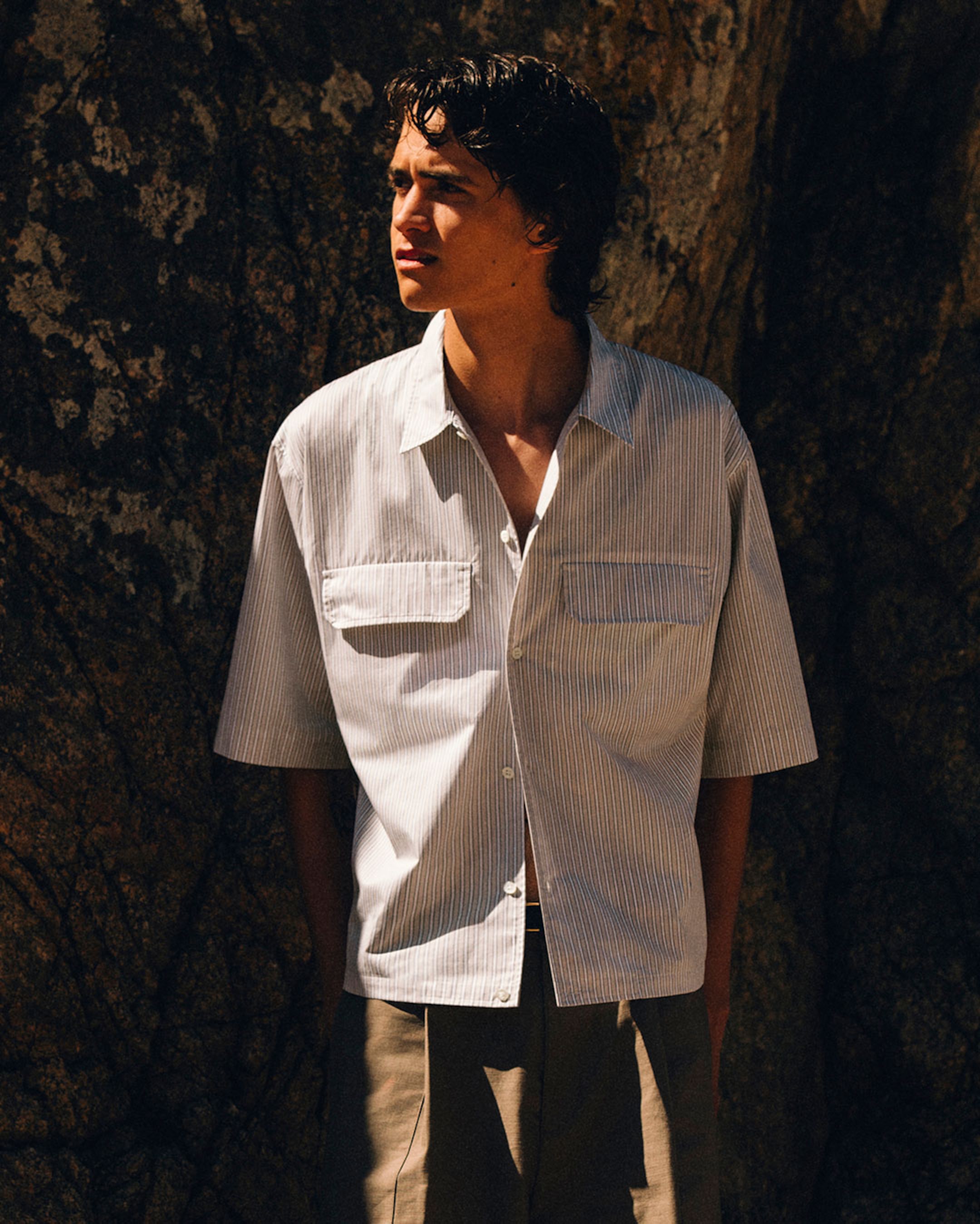 Young man in a light, short-sleeve striped shirt and khaki pants standing against a sunlit rock, looking to the left.