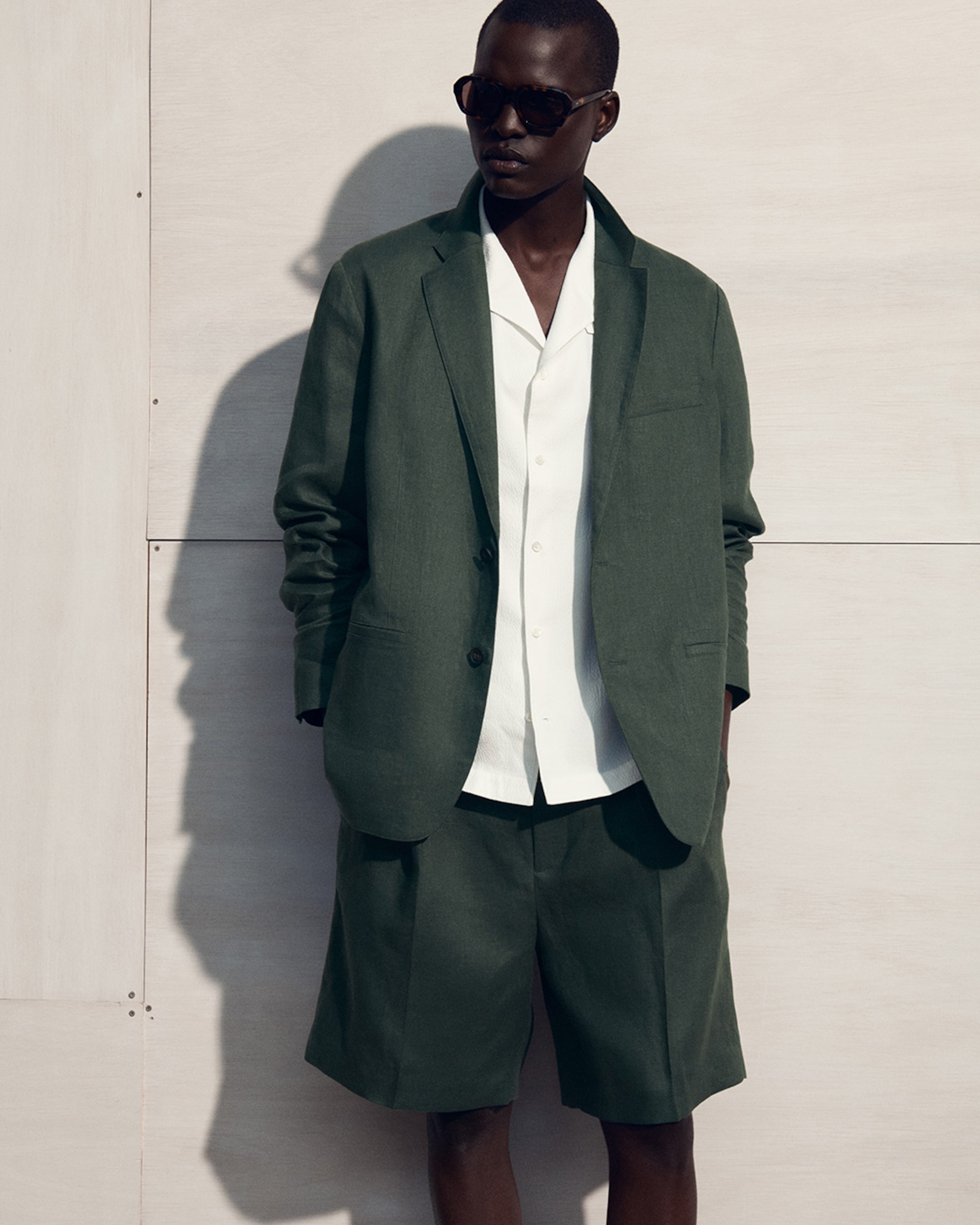 Person wearing a green suit jacket and shorts with a white shirt, standing against a light background, wearing sunglasses.
