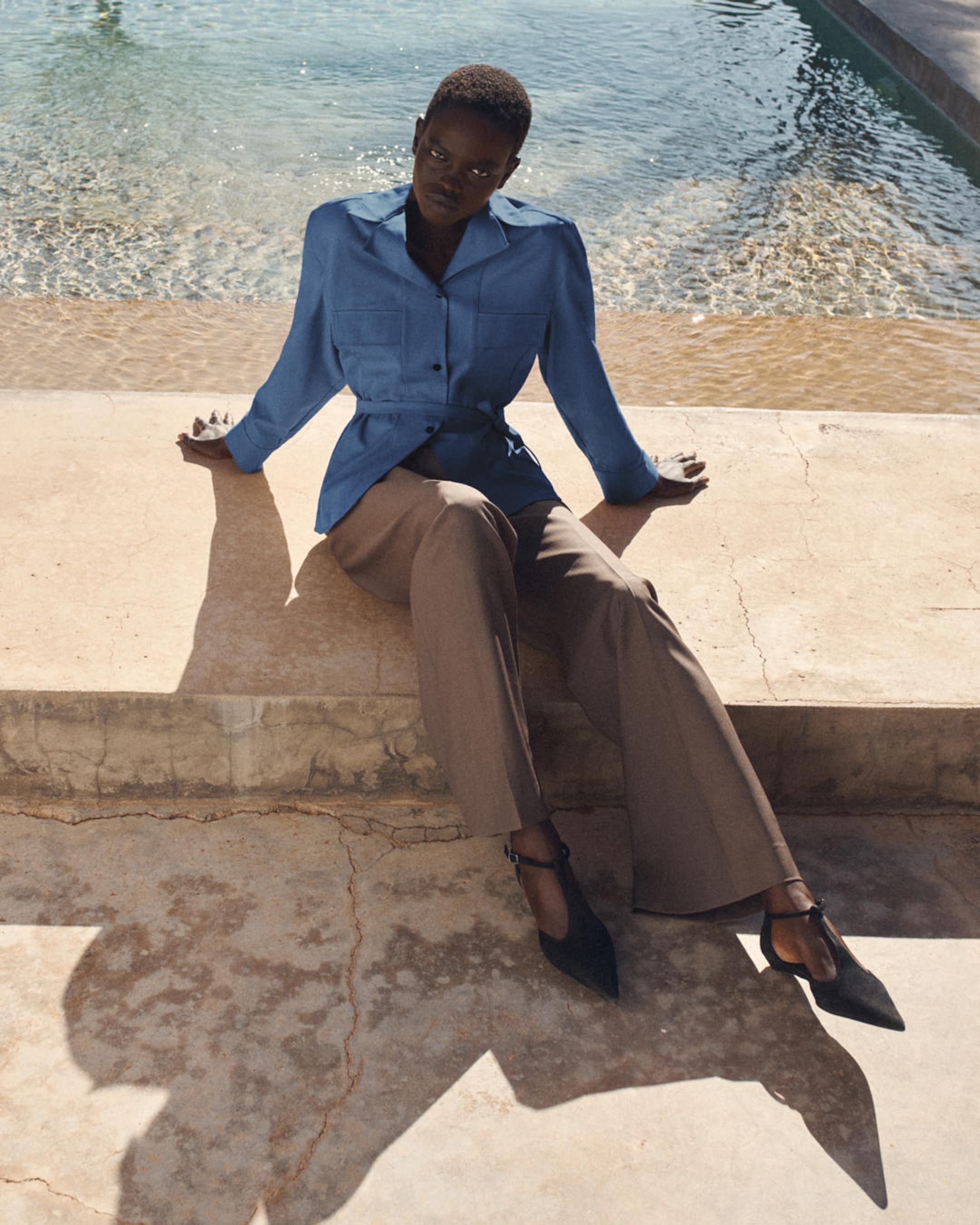 Person sitting on a stone ledge by a pool, wearing a blue blouse and brown trousers, with chic black shoes.