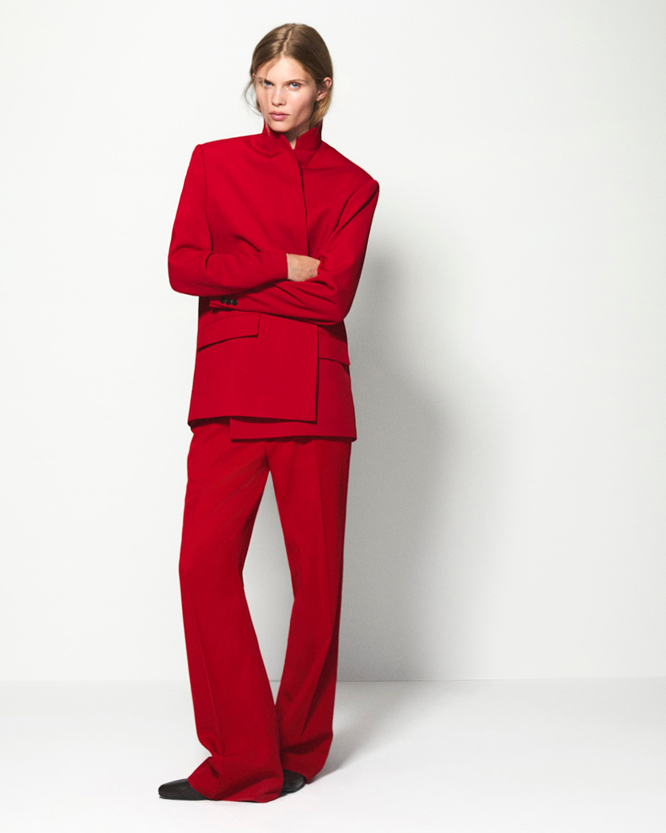 Person in a red tailored suit poses confidently against a plain backdrop, with a sleek hairstyle and black shoes.