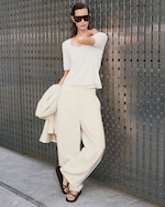 Placeholder for: A woman in beige pants and a white top leans against a textured metal wall, wearing sunglasses and black sandals, holding a coat.